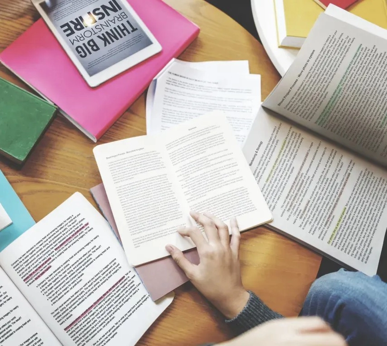 BD Medical Writing & Consulting - Draufsicht auf einen Holzschreibtisch: Offene Bücher, ein Tablet und verstreute Papiere liegen herum. Eine Hand hält ein Buch offen.