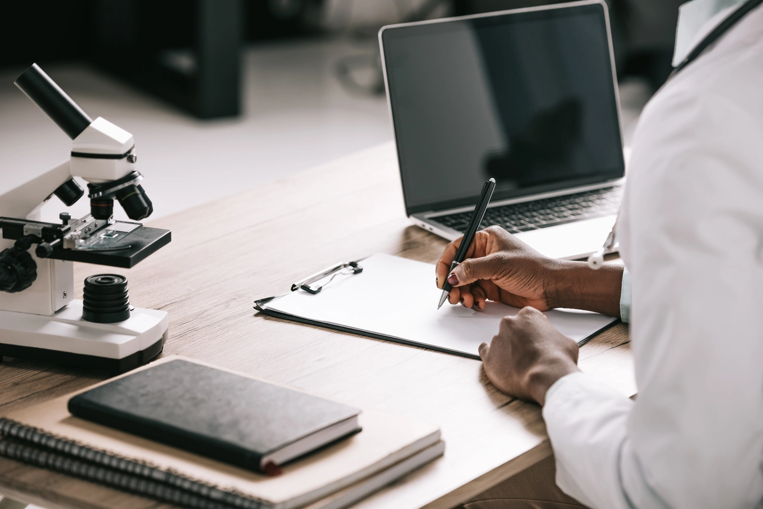 BD Medical Writing & Consulting - Ausschnitt eines afroamerikanischen Wissenschaftlers, der neben einem Laptop mit leerem Bildschirm auf Papier schreibt.