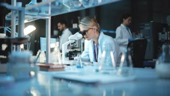 BD Medical Writing & Consulting - Eine Wissenschaftlerin im weißen Laborkittel arbeitet in einem modernen biomedizinischen Forschungslabor unter einem Mikroskop. Im Hintergrund sind Kolleginnen und Kollegen zu sehen.