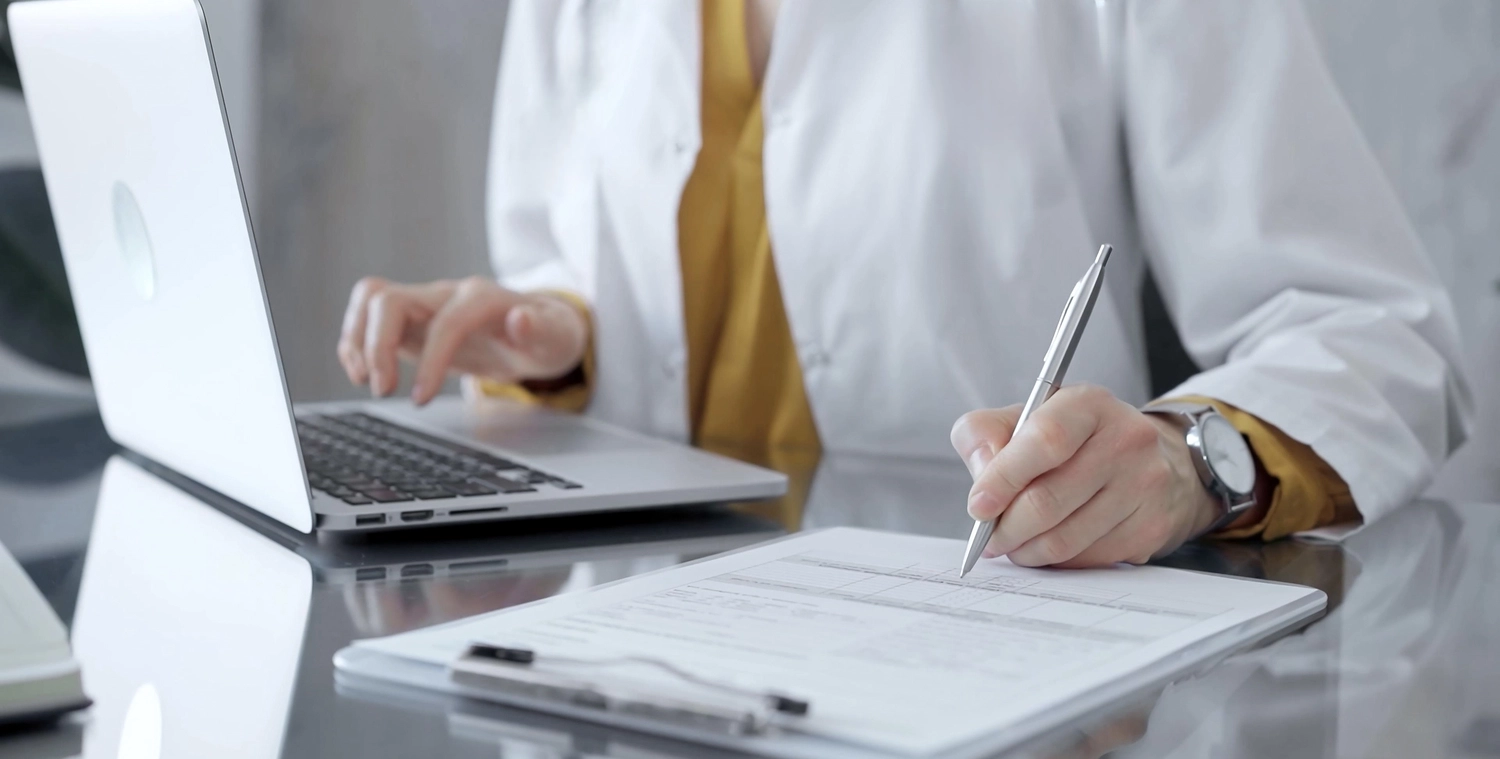 BD Medical Writing & Consulting - Eine Ärztin schreibt auf einem Glastisch an einem Dokument. Laptop und Tablet sind im Bild zu sehen. Arbeitsplatzkonzept im medizinischen Bereich.