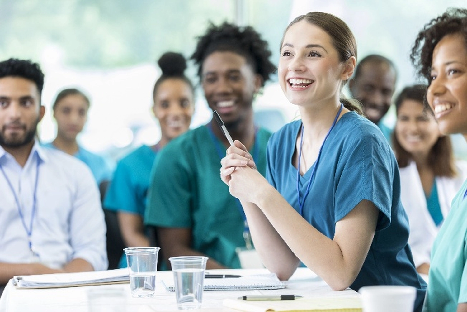 BD Medical Writing & Consulting - Eine lächelnde Krankenschwester in blauer Arbeitskleidung hält einen Stift und spricht während eines medizinischen Seminars oder einer Besprechung, umgeben von Kollegen unterschiedlicher Herkunft.